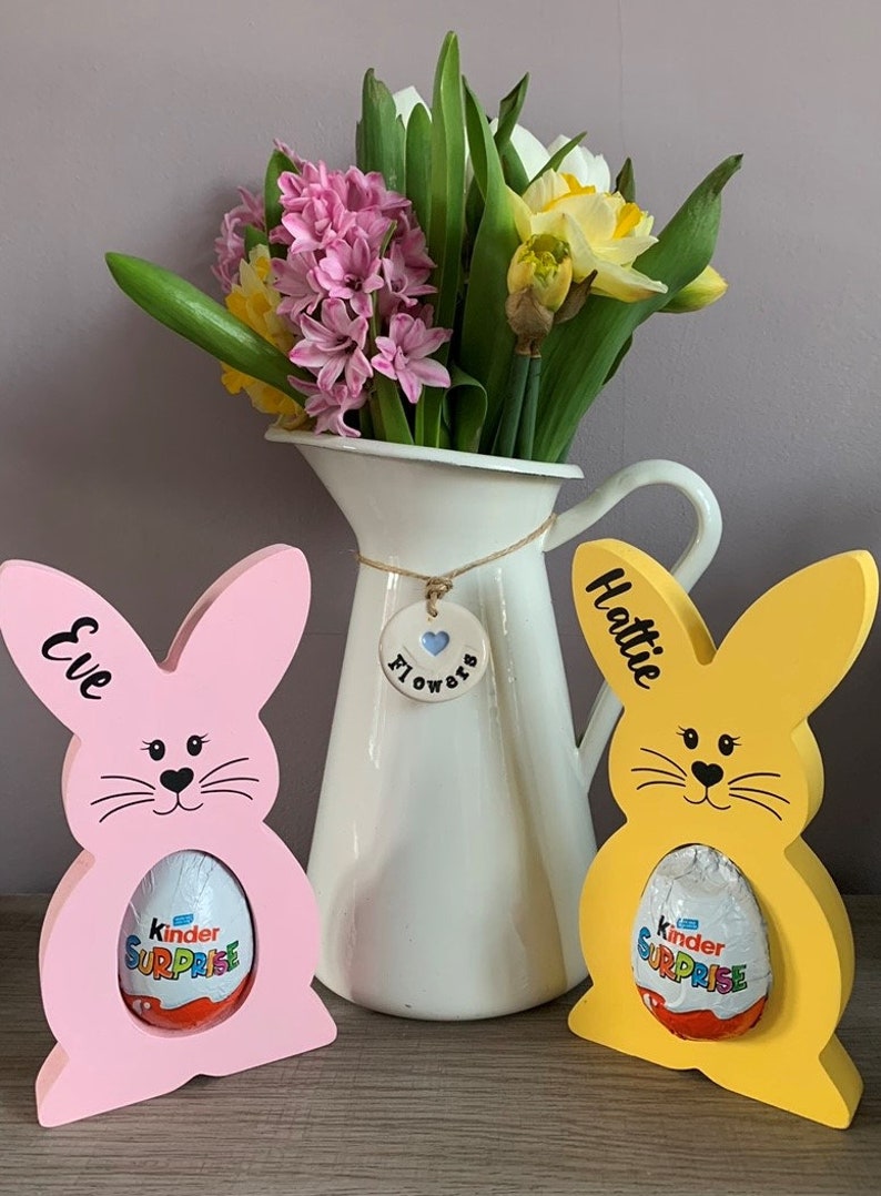 May include: Two wooden bunny shaped Easter egg holders, one pink and one yellow, with the names "Eve" and "Hattie" written on them. Each bunny is holding a Kinder Surprise chocolate egg. A white ceramic pitcher with a blue heart charm that says "Flowers" is in the background.
