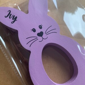 May include: Purple wooden bunny rabbit egg holder with a black face and whiskers. The bunny has a hole in the middle for an egg. The bunny has the name "Ivy" written on it.