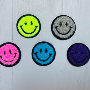 May include: Six embroidered smiley face patches in different colours: neon yellow, pink, light blue, purple, and grey. The patches are all the same size and shape, and they have a black outline.