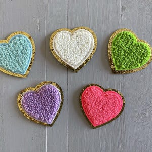 May include: Five heart-shaped felt patches with gold trim. The patches are in various colours: light blue, white, green, purple, and pink.