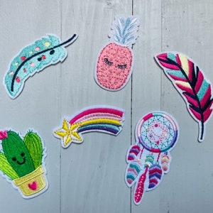 May include: A set of six embroidered iron-on patches featuring a variety of designs, including a pineapple, a cactus, a rainbow, a feather, a dreamcatcher, and a feather. The patches are all in bright colours and have a whimsical, boho style.