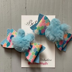 May include: Two blue and white hair bows with a fluffy blue pom-pom on each bow. The bows have a colourful, glittery pattern. The bows are on a white card with the text "Bettsickle Gifts" and "Derbyshire" written on it.