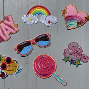 May include: A set of nine iron-on patches featuring various designs, including a rainbow, a heart with a rainbow inside, sunglasses, a pineapple, a lollipop, and the words "Yay" and "Stars".