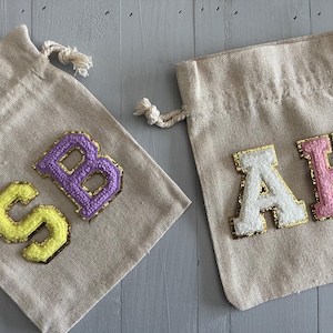 May include: Two small, beige fabric drawstring bags with felt letters sewn on. The bag on the left has the letters "SB" in yellow and purple. The bag on the right has the letters "AH" in white and pink.