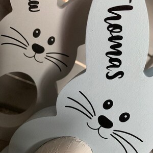 May include: Two light blue wooden bunny rabbit egg holders with black painted eyes, noses, and whiskers. The bunny on the left has the name "Matthew" written on it. The bunny on the right has the name "Thomas" written on it. Each bunny has a white chocolate egg inside.
