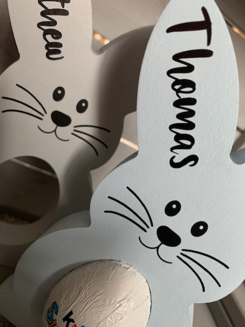May include: Two light blue wooden bunny rabbit egg holders with black painted eyes, noses, and whiskers. The bunny on the left has the name "Matthew" written on it. The bunny on the right has the name "Thomas" written on it. Each bunny has a white chocolate egg inside.