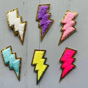 May include: Six lightning bolt shaped patches with gold outlines. The patches are made of a fuzzy material and come in the colours white, purple, pink, light blue, yellow, and hot pink.