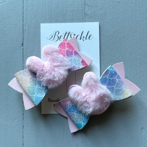 May include: Two pink and blue mermaid-themed hair bows with pink fluffy centres. The bows are handmade and have a glittery finish. The card in the background says "Bettsickle Handmade".