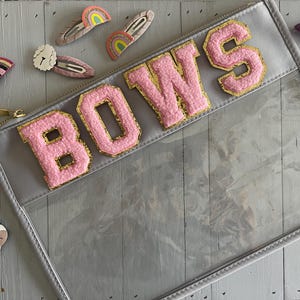May include: A grey, clear cosmetic bag with the word "BOWS" in pink, gold-trimmed letters. Hair clips in various designs, including rainbows and stars, are scattered around the bag. The bag has a gold zip.