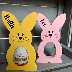May include: Two wooden bunny shaped Easter egg holders, one yellow and one pink. The yellow bunny has the name Hattie written on it and is holding a Kinder Surprise egg. The pink bunny has the name Eve written on it.