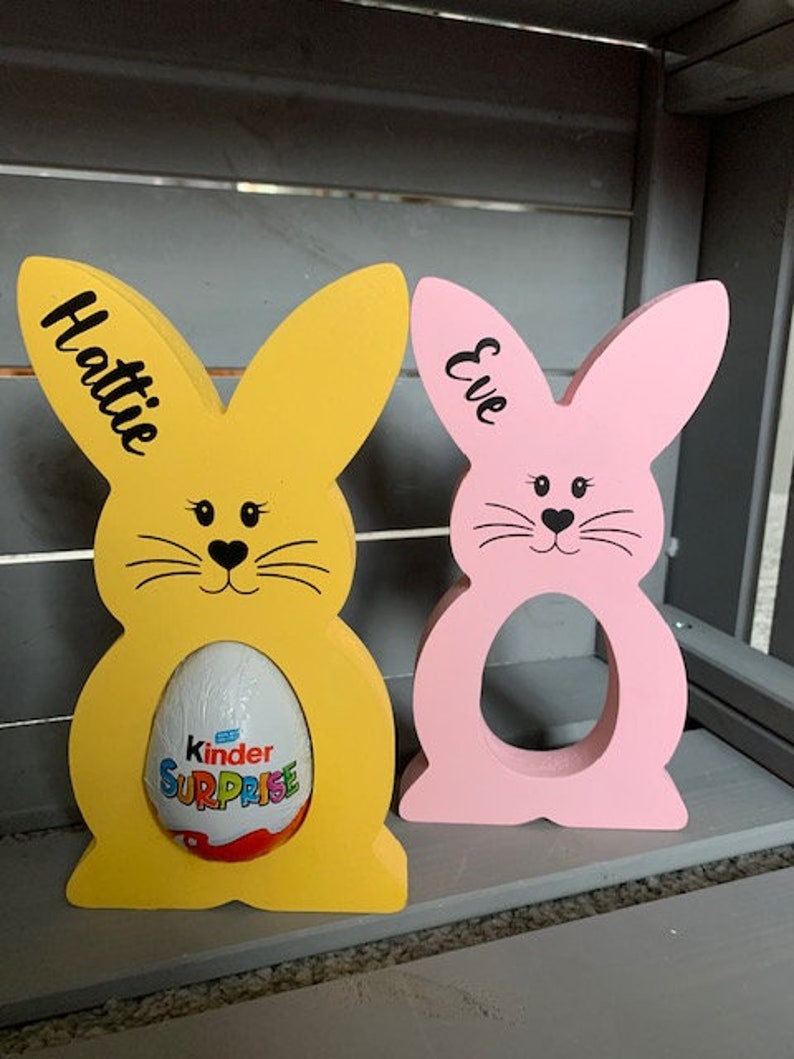 May include: Two wooden bunny shaped Easter egg holders, one yellow and one pink. The yellow bunny has the name Hattie written on it and is holding a Kinder Surprise egg. The pink bunny has the name Eve written on it.