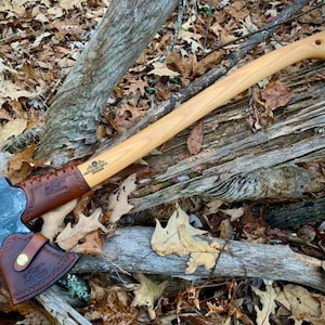 May include: A brown leather axe sheath with a brown leather strap and a brass button. The sheath is attached to a wooden-handled axe with a black metal head. The axe is lying on a bed of brown leaves and branches.
