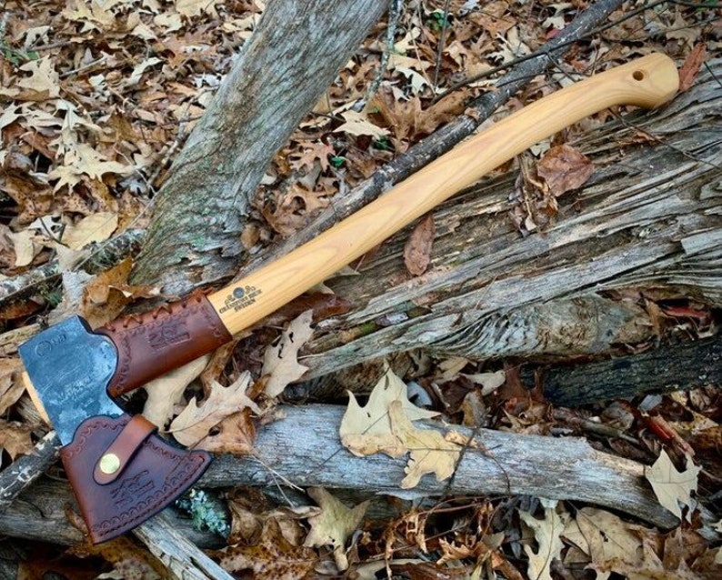 May include: A brown leather axe sheath with a brown leather strap and a brass button. The sheath is attached to a wooden-handled axe with a black metal head. The axe is lying on a bed of brown leaves and branches.