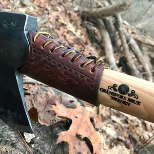 May include: A close-up of a Gränsfors Bruk axe with a leather handle wrap. The axe head is black and the handle is made of wood. The axe is made in Sweden.