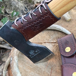 May include: A brown leather axe sheath with a  brass button and a  stitched design. The axe head is black with a  brown leather handle and a  stitched design. The axe handle is made of wood. The axe is on a  wooden surface.