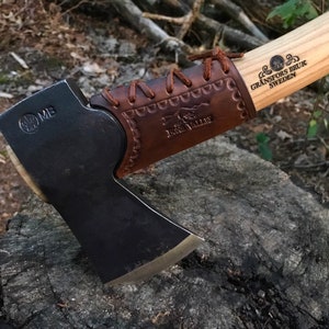May include: A brown leather-wrapped axe handle with a wood shaft. The axe head is black with a silver blade. The handle is stamped with "GRANSFORS BRUK SWEDEN" and "FOREST VALLEY".