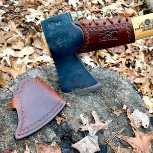 May include: A brown leather axe sheath with a decorative stitching pattern. The sheath is lying on a tree stump with brown leaves scattered around it. A black axe with a brown leather handle is visible in the background.