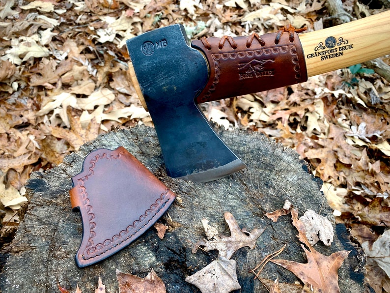 May include: A brown leather axe sheath with a decorative stitching pattern. The sheath is lying on a tree stump with brown leaves scattered around it. A black axe with a brown leather handle is visible in the background.