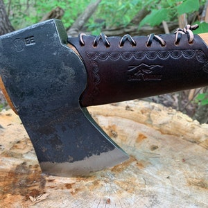May include: A black axe head with a brown leather handle wrap. The handle wrap has a decorative pattern and the text "Lone Wolf" is embossed on it. The axe is resting on a tree stump.