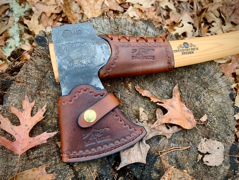 May include: A brown leather axe sheath with a stitched design and a brass button. The sheath is attached to a wooden-handled axe with a metal head. The axe handle is labeled "Gr&auml;nsfors Bruk Sweden".