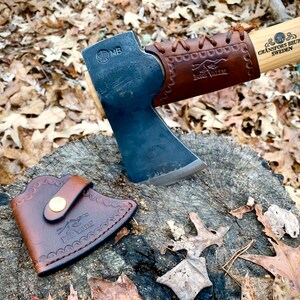 May include: A black axe with a brown leather handle and sheath. The axe head has the text "MB" stamped on it. The handle has the text "GRANSFORS BRUK SWEDEN" and "HULTQVIST" on it.