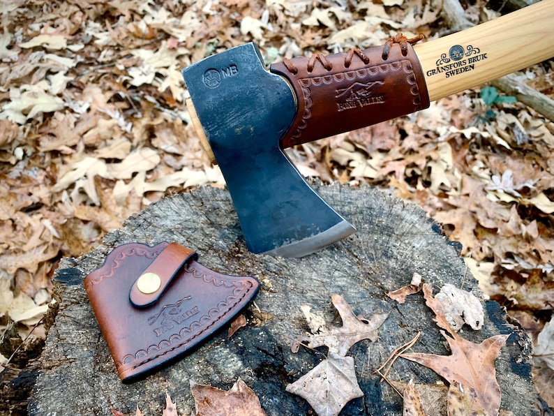May include: A black axe with a brown leather handle and sheath. The axe head has the text "MB" stamped on it. The handle has the text "GRANSFORS BRUK SWEDEN" and "HULTQVIST" on it.