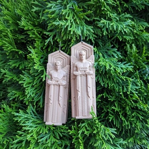May include: Two beige-colored ornaments shaped like a standing figure with a crown and intricate details. The ornaments are hanging from a green leafy branch.
