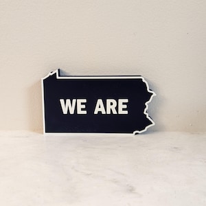 May include: A black and white outline of the state of Pennsylvania with the words "WE ARE" in white text on a dark blue background.