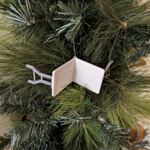K&ouml;nnte beinhalten: Wei&szlig;er Weihnachtsbaumschmuck in Form eines Klappstuhls. Der Schmuck h&auml;ngt an einem gr&uuml;nen k&uuml;nstlichen Weihnachtsbaum.