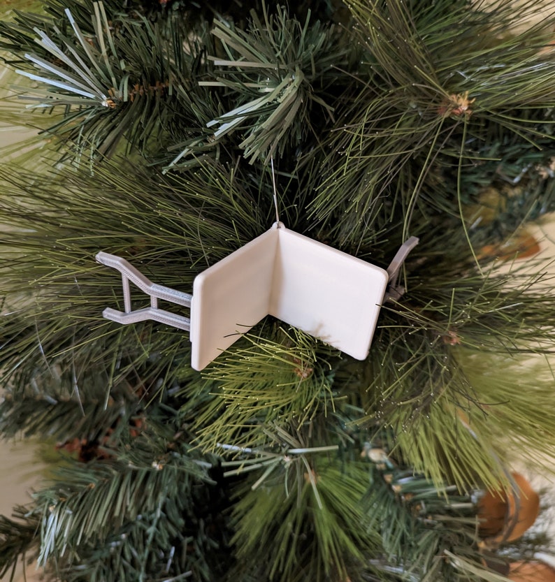 K&ouml;nnte beinhalten: Wei&szlig;er Weihnachtsbaumschmuck in Form eines Klappstuhls. Der Schmuck h&auml;ngt an einem gr&uuml;nen k&uuml;nstlichen Weihnachtsbaum.