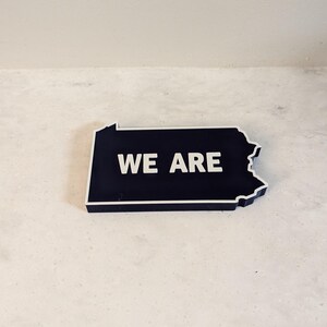 May include: A black outline of the state of Pennsylvania with the words "WE ARE" in white lettering.