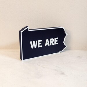May include: A dark blue wooden sign shaped like the state of Pennsylvania with the words "WE ARE" in white lettering.