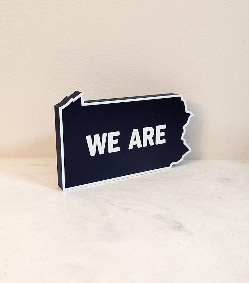 May include: A dark blue wooden sign shaped like the state of Pennsylvania with the words "WE ARE" in white lettering.