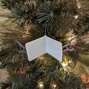 K&ouml;nnte beinhalten: Ein wei&szlig;er, gefalteter, buchf&ouml;rmiger Weihnachtsbaumschmuck mit einem grauen Metallhalter. Der Schmuck h&auml;ngt an einem Weihnachtsbaum.