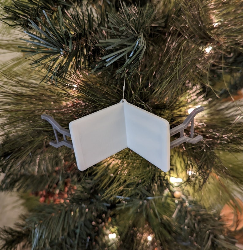 K&ouml;nnte beinhalten: Ein wei&szlig;er, gefalteter, buchf&ouml;rmiger Weihnachtsbaumschmuck mit einem grauen Metallhalter. Der Schmuck h&auml;ngt an einem Weihnachtsbaum.