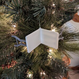 K&ouml;nnte beinhalten: Wei&szlig;er Weihnachtsbaumschmuck in Form eines Buches mit einem grauen Metallhalter. Der Schmuck ist leer und kann personalisiert werden.