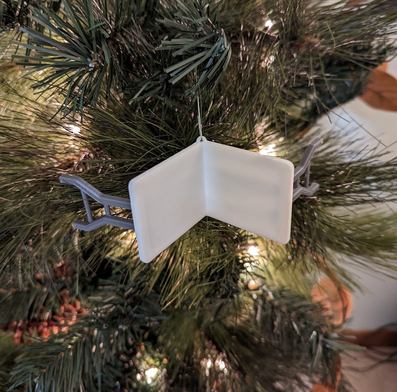 K&ouml;nnte beinhalten: Wei&szlig;er Weihnachtsbaumschmuck in Form eines Buches mit einem grauen Metallhalter. Der Schmuck ist leer und kann personalisiert werden.
