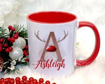 May include: White ceramic mug with a red rim and handle. The mug features a watercolor illustration of a reindeer with a red nose and the letter "A" in gold. The name "Ashleigh" is written in red cursive below the reindeer.