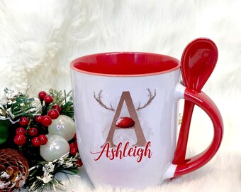 May include: White ceramic mug with a red rim and handle. The mug features a watercolor illustration of a reindeer with antlers and the letter "A" in gold. The name "Ashleigh" is written in cursive below the illustration. The mug comes with a matching red spoon.