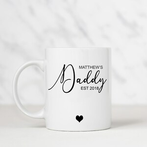 May include: White ceramic mug with a black inscription that reads "Matthew's Daddy Est 2018" and a small black heart.