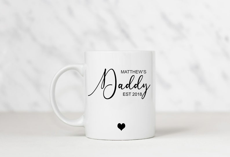 May include: White ceramic mug with a black inscription that reads "Matthew's Daddy Est 2018" and a small black heart.