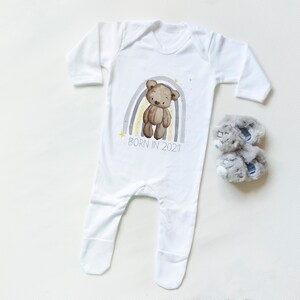May include: White baby onesie with a brown teddy bear standing on a rainbow with yellow and gray stripes. The text "BORN IN 2021" is printed below the rainbow. A pair of gray baby booties are also pictured.