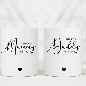 May include: Two white ceramic mugs with black text and a small black heart on the bottom. The mug on the left says "Perry's Mummy Est 2018". The mug on the right says "Perry's Daddy Est 2018".