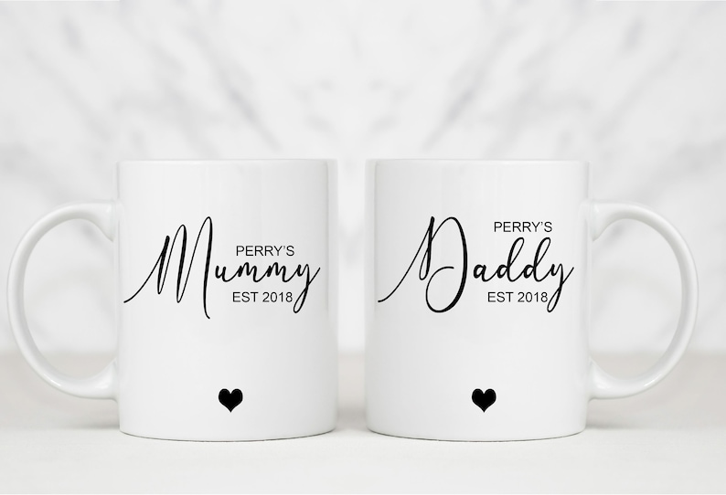 May include: Two white ceramic mugs with black text and a small black heart on the bottom. The mug on the left says "Perry's Mummy Est 2018". The mug on the right says "Perry's Daddy Est 2018".