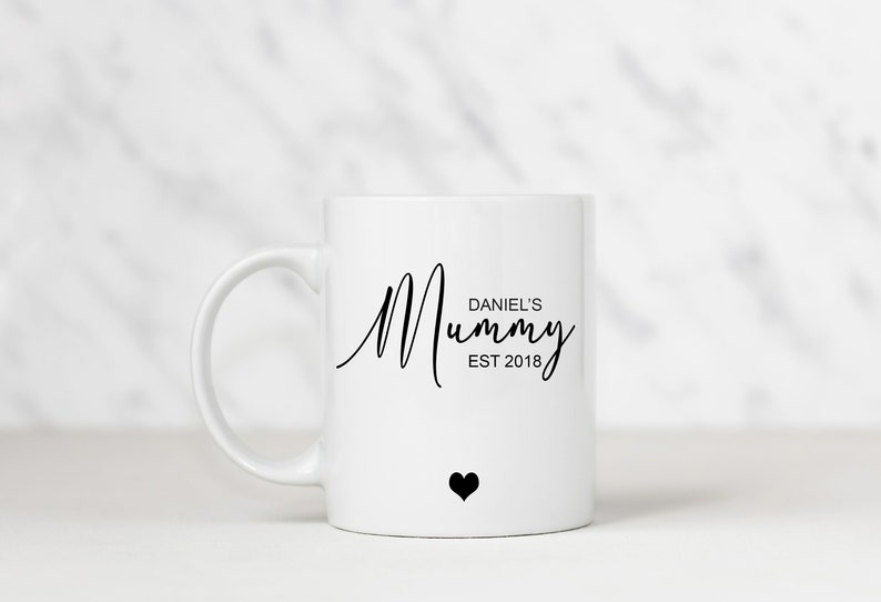 May include: White ceramic mug with a black inscription that reads "Daniel's Mummy Est 2018" and a small black heart.