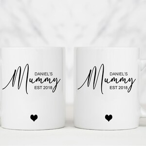May include: Two white ceramic mugs with black handles. Both mugs have the text "Mummy" in a cursive font with the words "Daniel's" and "EST 2018" underneath. Each mug has a small black heart at the bottom.