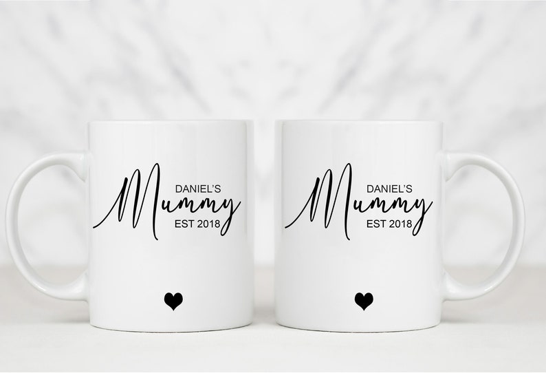 May include: Two white ceramic mugs with black handles. Both mugs have the text "Mummy" in a cursive font with the words "Daniel's" and "EST 2018" underneath. Each mug has a small black heart at the bottom.