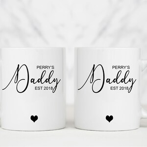 May include: Two white ceramic mugs with black text that reads "Perry's Daddy Est 2018" and a small black heart on the bottom of each mug.