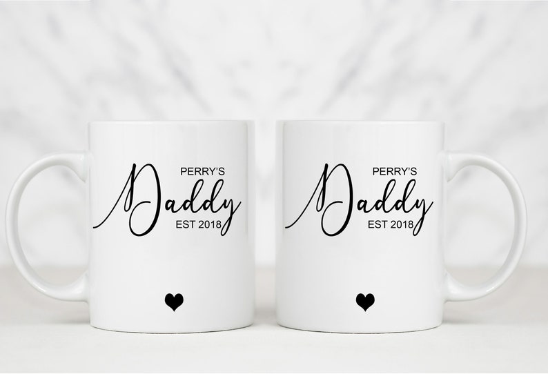 May include: Two white ceramic mugs with black text that reads "Perry's Daddy Est 2018" and a small black heart on the bottom of each mug.