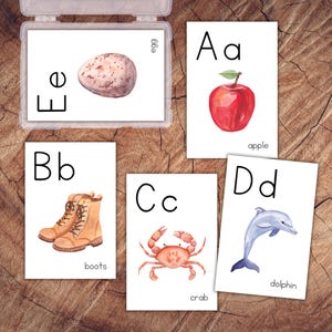 May include: A set of five educational alphabet flashcards featuring watercolour illustrations and black text. Cards display A for apple, B for boots, C for crab, D for dolphin, and E for egg. One card is inside a clear plastic storage case.